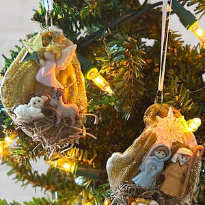 Può includere: Due decorazioni natalizie appese a un albero. Un ornamento presenta un angelo, un agnello e un asino. L'altro raffigura Maria, Giuseppe e Gesù bambino. Entrambi gli ornamenti hanno una finitura glitterata dorata.