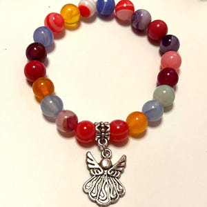 Pode incluir: Uma pulseira de contas coloridas com um pingente de anjo prateado. A pulseira apresenta contas redondas em tons de vermelho, laranja, amarelo, azul e roxo. O pingente de anjo tem asas e vestes esvoa&ccedil;antes.