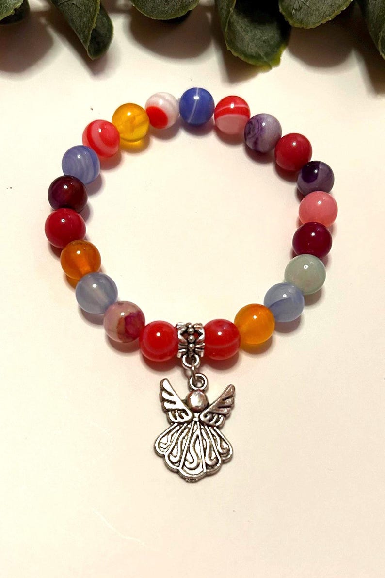 Pode incluir: Uma pulseira de contas coloridas com um pingente de anjo prateado. A pulseira apresenta contas redondas em tons de vermelho, laranja, amarelo, azul e roxo. O pingente de anjo tem asas e vestes esvoa&ccedil;antes.