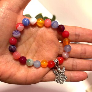 Pode incluir: Uma pulseira colorida de contas com um berloque de anjo prateado. A pulseira apresenta uma variedade de contas redondas em tons de vermelho, rosa, roxo, azul, amarelo e laranja. O berloque de anjo &eacute; detalhado com asas e um vestido esvoa&ccedil;ante.