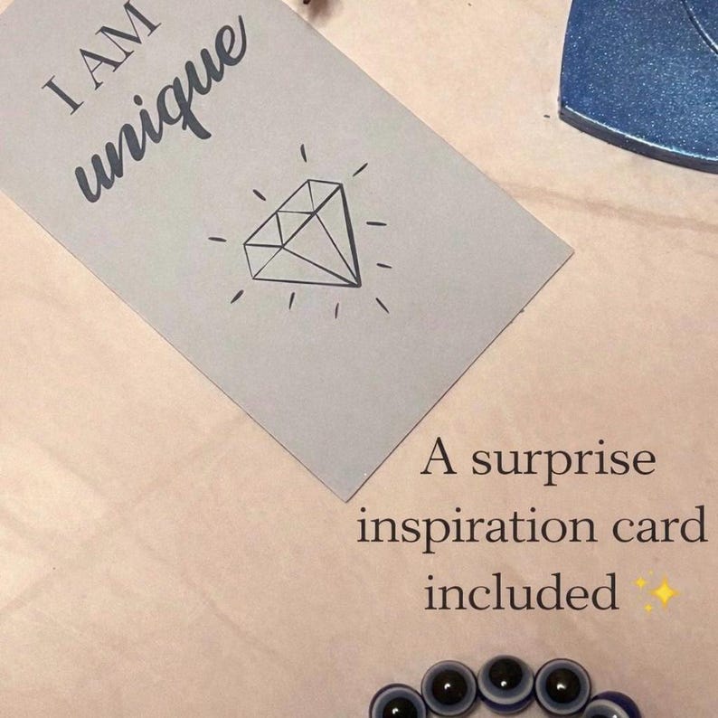 May include: A light gray card with the words "I AM unique" and a diamond illustration. The card is accompanied by a surprise inspiration card. A blue and black beaded bracelet is visible at the bottom.