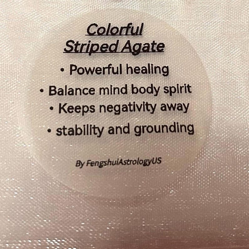 Pode incluir: Um adesivo circular branco com o texto "Colorful Striped Agate" e marcadores que listam benef&iacute;cios como "Cura poderosa" e "Equil&iacute;brio mente corpo esp&iacute;rito". O adesivo est&aacute; sobre um fundo texturizado esbranqui&ccedil;ado.