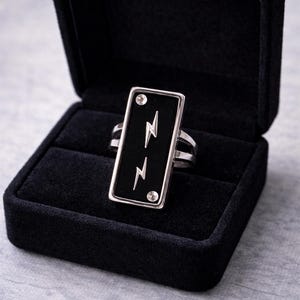 May include: A silver ring with a rectangular black onyx stone featuring two lightning bolt designs. The ring is set in a black velvet jewelry box. Two small, round, clear stones are set at the top and bottom of the black onyx stone.