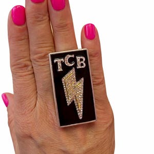 May include: A rectangular ring featuring a black enamel face with the letters TCB in silver-tone metal above a lightning bolt motif. The letters and the lightning bolt are encrusted with small clear crystals, and the ring has a silver-tone metal border.