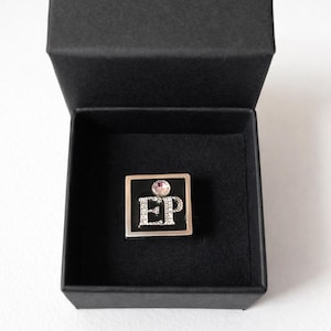 May include: A silver ring with a black square face featuring the letters "EP" in a sparkling, silver font. A small, round, pink gemstone sits above the letters. The ring is presented in an open, black jewelry box.
