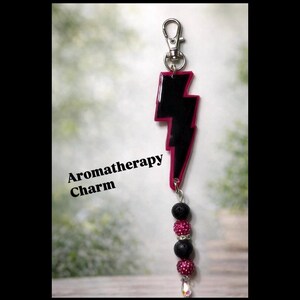 May include: Aromatherapy charm with a black and pink lightning bolt design, a silver clasp, and a string of beads. The beads include black lava stones, pink rhinestone beads, and a clear crystal charm. The text "Aromatherapy Charm" is visible.