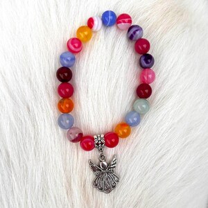 Pode incluir: Uma pulseira de contas coloridas com um pingente de anjo prateado. A pulseira apresenta contas redondas multicoloridas, incluindo vermelho, laranja, amarelo, azul e roxo. Um pingente de anjo prateado detalhado pende dela.