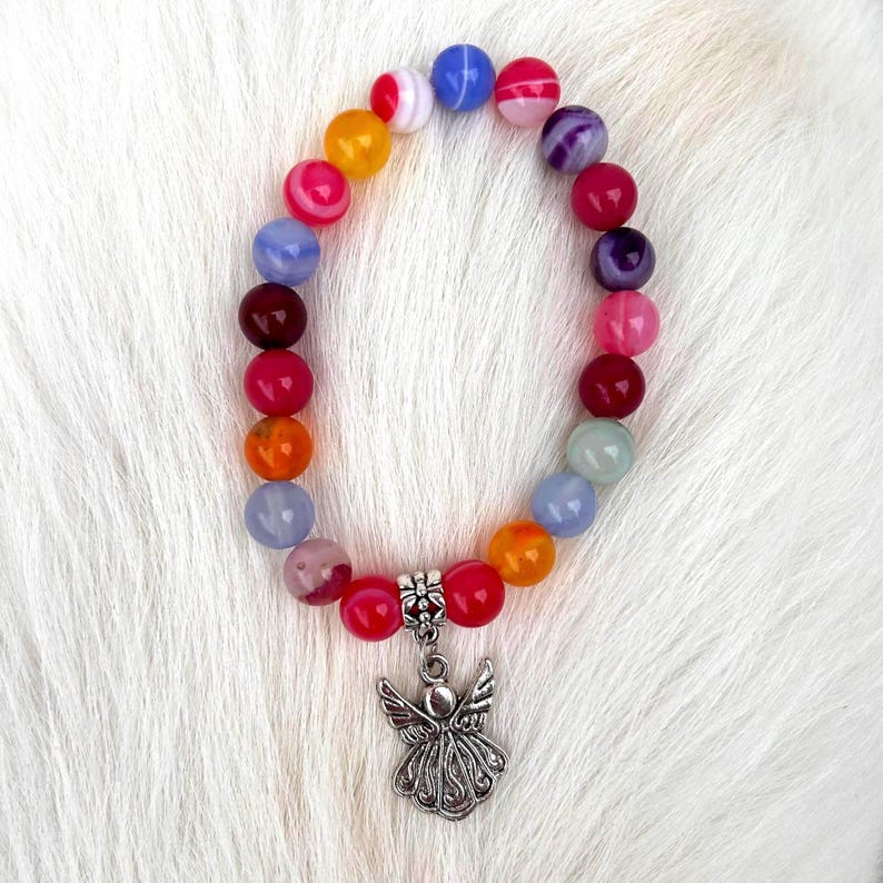 Pode incluir: Uma pulseira de contas coloridas com um pingente de anjo prateado. A pulseira apresenta contas redondas multicoloridas, incluindo vermelho, laranja, amarelo, azul e roxo. Um pingente de anjo prateado detalhado pende dela.