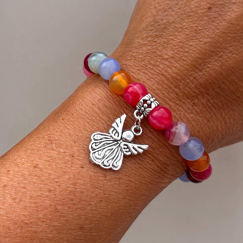 Pode incluir: Uma pulseira de contas com pedras preciosas redondas coloridas em tons de rosa, laranja, azul e roxo. A pulseira inclui um berloque de anjo prateado. A pulseira &eacute; usada no pulso.