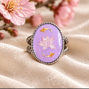 May include: A silver-toned ring featuring an oval-shaped lavender resin inlay. The center of the inlay displays a pink lotus flower graphic flanked by two small orange and white koi fish. The ring band has a twisted, rope-like metal texture.