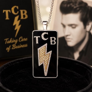 May include: A rectangular silver-tone pendant necklace featuring a black enamel inlay. The pendant displays the letters T, C, and B in clear crystals, with a central lightning bolt motif filled with yellow and clear crystals on a silver chain.