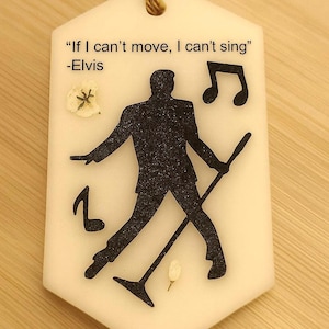 May include: A white, hexagonal ornament with a gold string for hanging. The ornament features a black silhouette of a singer with a microphone, musical notes, and the quote "If I can't move, I can't sing" by Elvis.