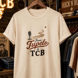 May include: A cream-colored short-sleeve t-shirt featuring a graphic of a microphone, a winding road, and a star. The text reads "From Tupelo to TCB" in brown and black lettering. The shirt has a crew neckline and rolled sleeve cuffs.
