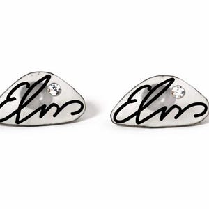 May include: A pair of silver-toned stud earrings. Each earring is a rounded, irregular shape with a small, clear crystal and the word "Elms" in black script. The earrings are set against a white background.