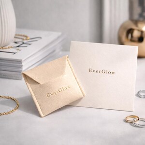 May include: A beige suede jewelry pouch and a matching card with the text "EverGlow" in gold lettering. The pouch is open, and the card is square. A gold chain and rings are visible.