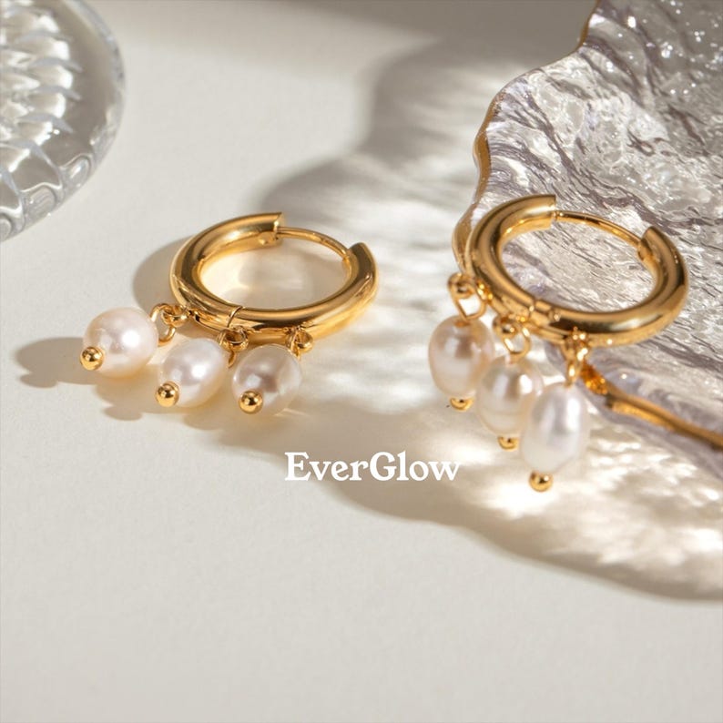 May include: Two gold hoop earrings with dangling white pearls. The earrings are displayed on a white surface with a clear, textured object in the background. The brand name "EverGlow" is printed in white.