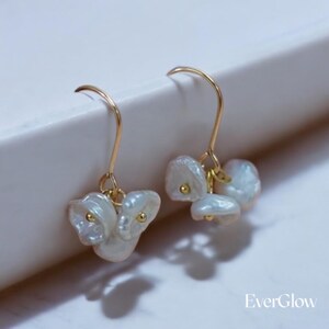 May include: A pair of gold dangle earrings with white pearl-like drops. The earrings have a simple design and are perfect for everyday wear.