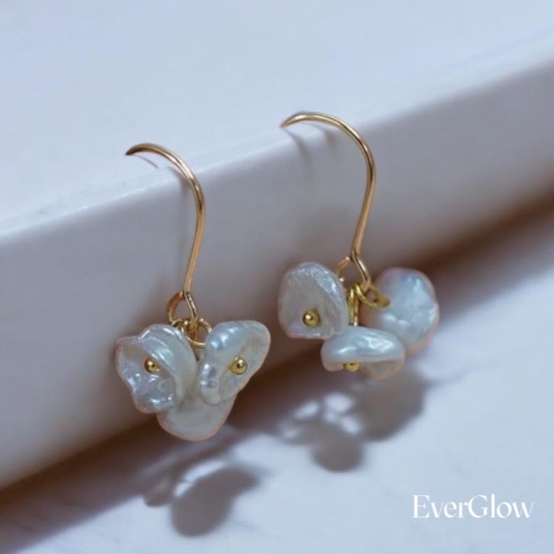 May include: A pair of gold dangle earrings with white pearl-like drops. The earrings have a simple design and are perfect for everyday wear.