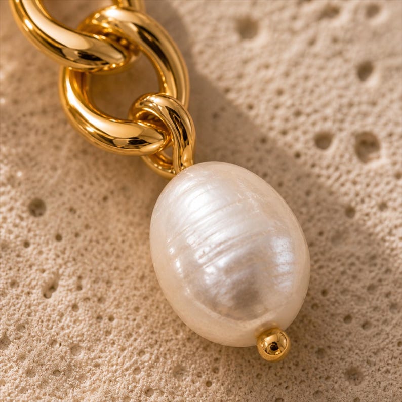May include: Close-up of a gold-colored chain link with a large, oval, white pearl pendant. The pearl has a lustrous surface and is attached to the chain with a small gold bead. The background is a textured, light beige surface.