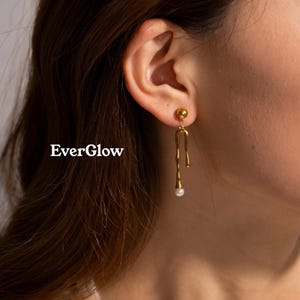 May include: Gold-colored drop earrings with a unique, flowing design. Each earring features a small, round stud at the top, a curved, elongated shape, and a small, white pearl at the bottom. The earrings are displayed near a person's ear. The text "EverGlow" is visible.