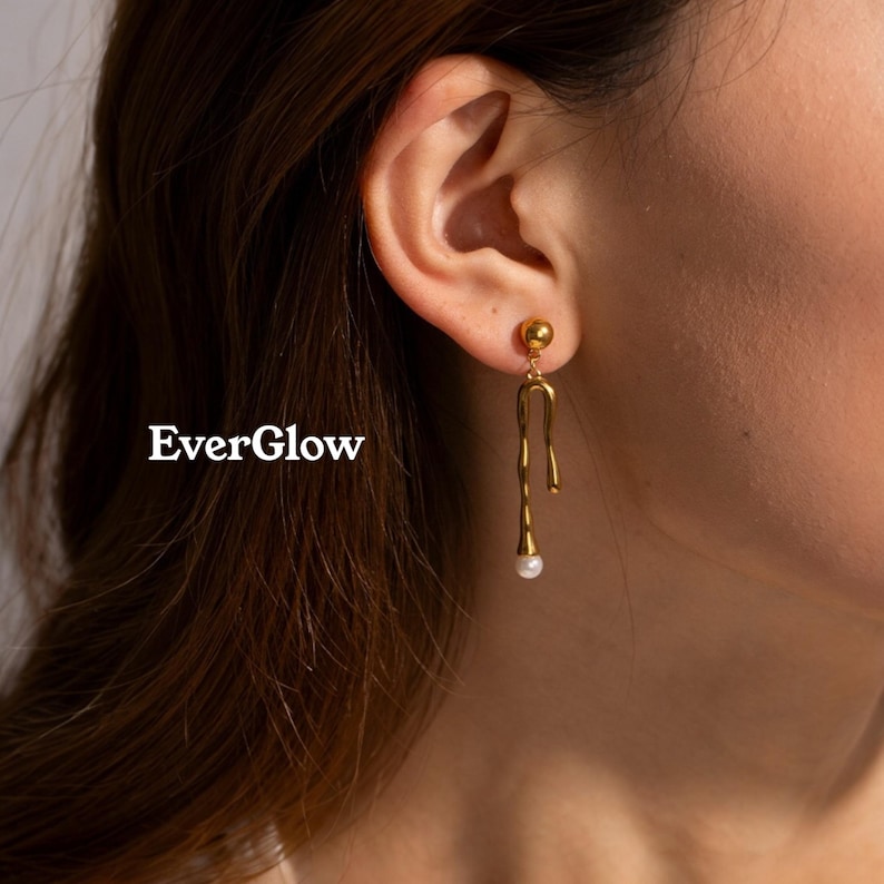 May include: Gold-colored drop earrings with a unique, flowing design. Each earring features a small, round stud at the top, a curved, elongated shape, and a small, white pearl at the bottom. The earrings are displayed near a person's ear. The text "EverGlow" is visible.