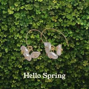 May include: A pair of gold hoop earrings with white pearl-like flower accents. The earrings are on a green background with the text "Hello Spring".