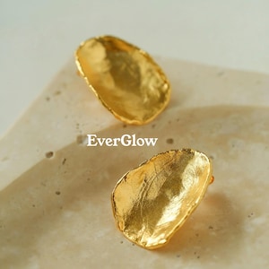 Może przedstawiać: Dwa złote kolczyki w kształcie nieregularnych, organicznych liści. Kolczyki leżą na jasnobeżowej powierzchni.