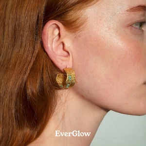 Puede incluir: Primer plano de un pendiente de aro texturizado de color dorado con pequeños detalles turquesas. El pendiente se lleva en la oreja de una persona con pelo rojo. El texto "EverGlow" está en la parte inferior de la imagen.