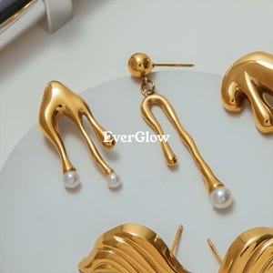 May include: Close-up of gold-colored earrings with a unique, flowing design. One pair features a teardrop shape with a pearl at the bottom, while another has a curved, elongated shape with a pearl. The brand name "EverGlow" is visible.