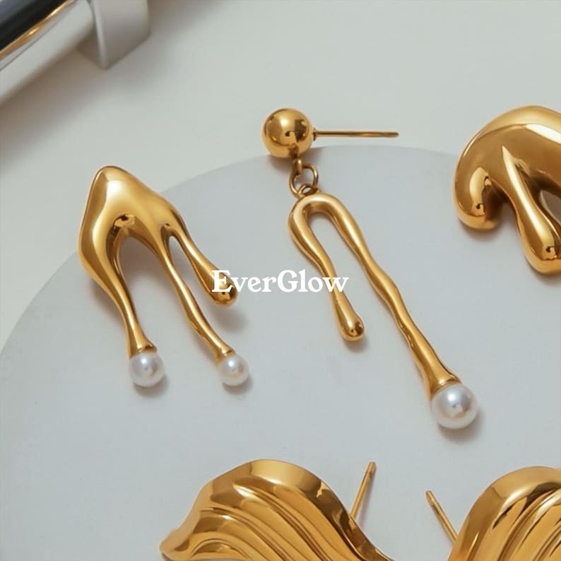 May include: Close-up of gold-colored earrings with a unique, flowing design. One pair features a teardrop shape with a pearl at the bottom, while another has a curved, elongated shape with a pearl. The brand name "EverGlow" is visible.