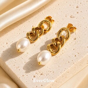 May include: A pair of gold-colored chain link earrings with a dangling white pearl. The earrings have a post back and are displayed on a beige surface. The brand name "EverGlow" is visible at the bottom.
