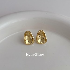 Peut inclure: Une paire de boucles d'oreilles dor&eacute;es avec une forme ovale irr&eacute;guli&egrave;re textur&eacute;e. Les boucles d'oreilles sont sur une surface blanche.