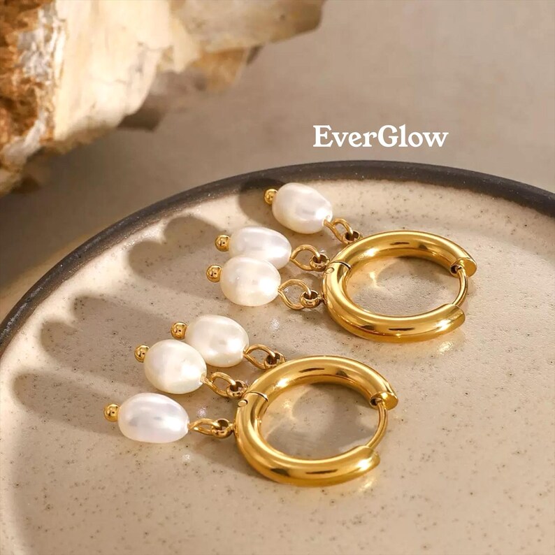 May include: A pair of gold-colored hoop earrings with dangling white pearls. The earrings are on a light-colored plate. The word "EverGlow" is printed in white.