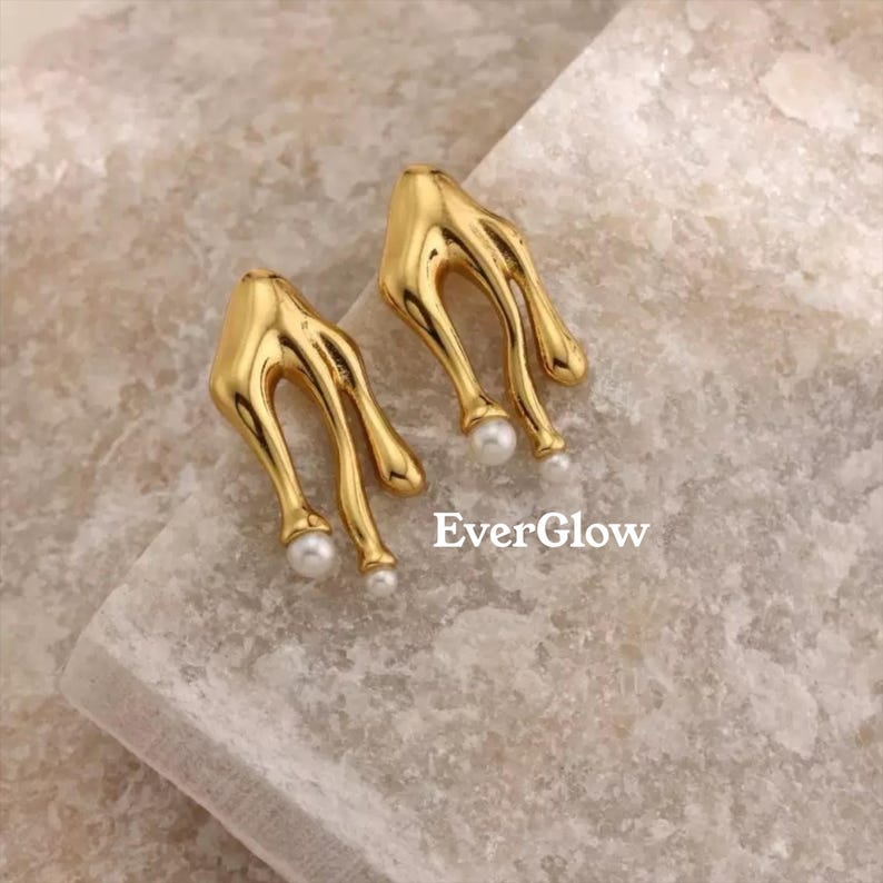 May include: A pair of gold-colored earrings with a unique, flowing design. Each earring features three curved, elongated elements, with a small, white pearl at the end of two of the elements. The text "EverGlow" is visible.