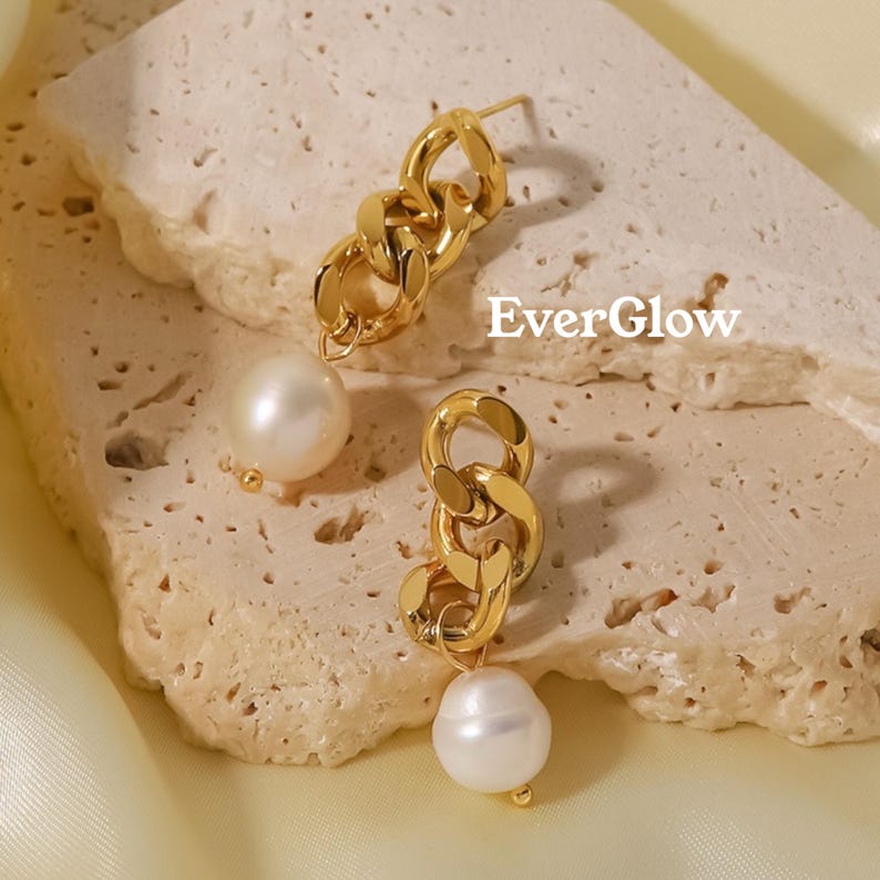 May include: A pair of gold-colored chain link earrings with a pearl drop. The earrings have a post back and are displayed on a textured, light-colored surface. The word "EverGlow" is printed on the surface.