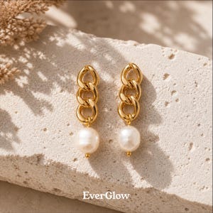 May include: A pair of gold-colored chain link earrings with a large, white pearl drop. The earrings are displayed on a light-colored stone surface. The brand name "EverGlow" is printed at the bottom.