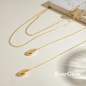 May include: Gold-colored layered necklaces with delicate chains and shell pendants. The necklaces are displayed on a light-colored surface, with the EverGlow logo in the bottom right corner. The chains are thin and the pendants are small.