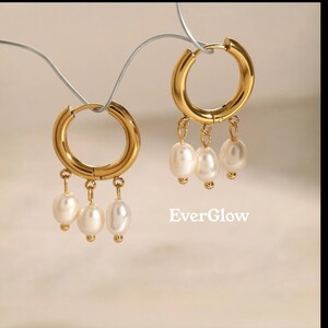 May include: Gold-colored hoop earrings with dangling white pearls. The earrings have a polished finish and a simple, elegant design. The image includes the text "EverGlow".