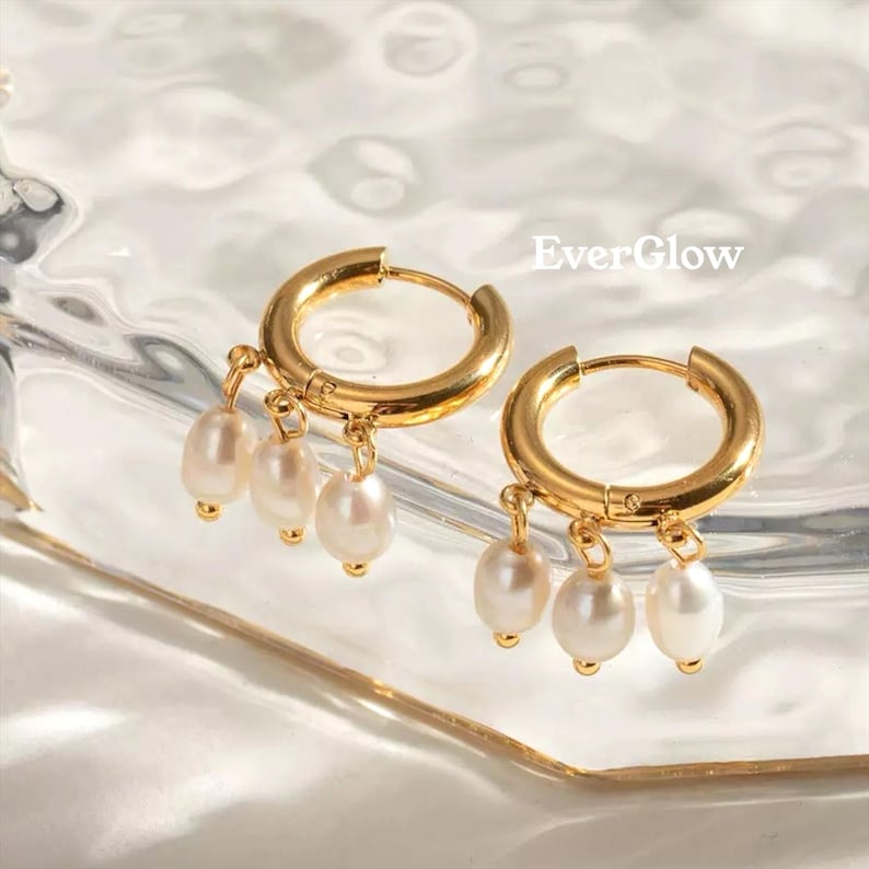 May include: Gold-colored hoop earrings with dangling white pearls. The earrings have a polished finish and are displayed on a reflective surface. The text "EverGlow" is visible in the image. These earrings are a jewelry accessory.