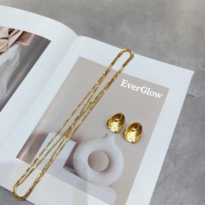 Peut inclure: Un collier en cha&icirc;ne dor&eacute;e et une paire de boucles d'oreilles dor&eacute;es &agrave; la texture martel&eacute;e. Les boucles d'oreilles sont de forme ovale. Le texte "EverGlow" est visible sur l'image.