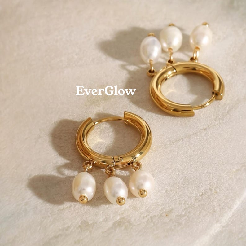 May include: Two gold-colored hoop earrings with dangling white pearls. The earrings are on a light-colored surface. The text "EverGlow" is in the upper left corner.