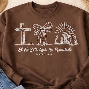 Może przedstawiać: Brązowa bluza z białym rysunkiem krzyża, kokardy i grobu. Tekst "Él No Está Aquí, Ha Resucitado" i "MATEO 28:6" są wydrukowane na biało. Bluza jest połączona z niebieskimi dżinsami i słomkowym kapeluszem.