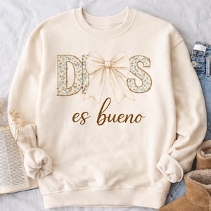 May include: Cream-colored sweatshirt with the words "DIOS es bueno" in brown script. The letters "DIOS" are filled with a floral pattern, and a large bow is centered above the text. The sweatshirt has long sleeves.