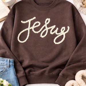 May include: A brown sweatshirt with the word "Jesus" spelled out in a cursive font using off-white, pearl-like beads. The sweatshirt is displayed with blue jeans, a cup of coffee, a candle, and slippers.