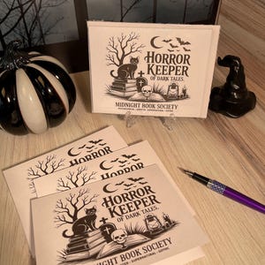 May include: White cards with the text "HORROR KEEPER OF DARK TALES" and a black cat illustration. A black and white pumpkin and a black witch's hat are also visible.