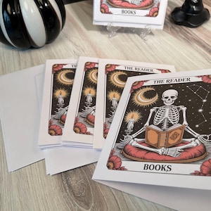 Peut inclure: Cartes de vœux avec un squelette lisant un livre, avec le texte "THE READER" et "BOOKS". Les cartes ont un design céleste avec un croissant de lune, des bougies et des motifs floraux. Comprend des enveloppes blanches.