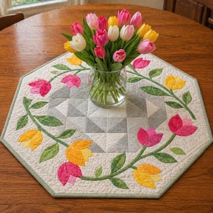 May include: A colorful bouquet of tulips in a clear glass vase sits on an octagonal quilted table runner. The runner features a floral design with pink and yellow tulips, green leaves, and a geometric center. The table is made of wood.