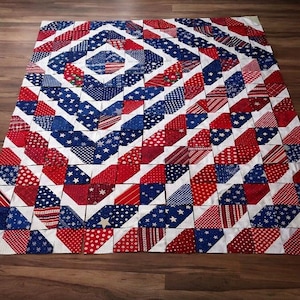 May include: A handmade quilt with a patriotic theme. The quilt features a geometric pattern of red, white, and blue fabrics, including stars and stripes. The quilt is laid out on a wooden surface.