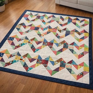 May include: A colorful quilt with a white background and a zig-zag pattern made of various fabric pieces in shades of red, blue, green, and yellow. The quilt has a dark blue border and is placed on a wooden floor.