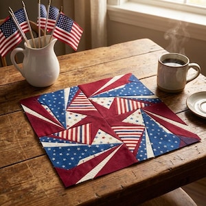 May include: A square fabric quilt block featuring a geometric star pattern in red, white, and blue. The design incorporates solid red fabric, blue fabric with white stars, and red and white striped fabric, arranged in a complex, angular, pieced layout.
